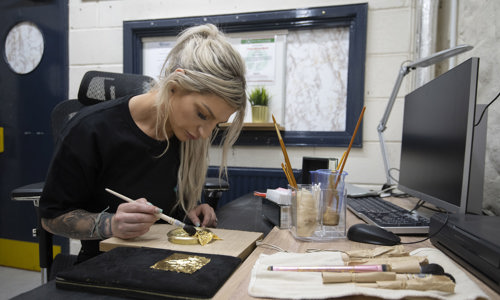 A woman sitting at a desk applying gold leaf to the Lion Rampant Roundels 