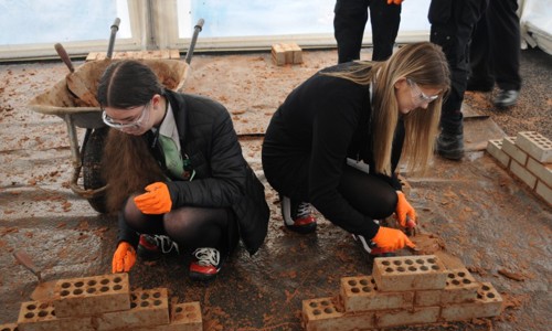 Two young people are laying bricks in a learning workshop. They both wear safety goggles and protective gloves.